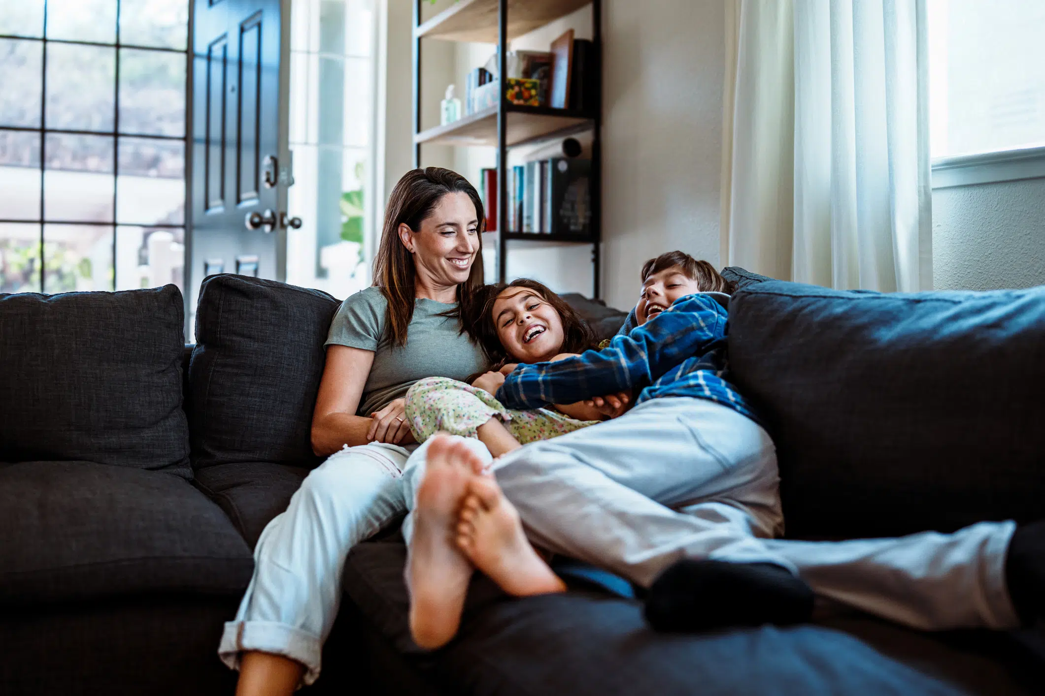 Elementary age brother and sister have a playful tickle fight with their mother on the sofa in the living room of their home.
